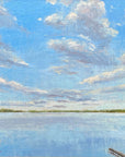 Framed painting of a blue sky with clouds over water on a white wall.